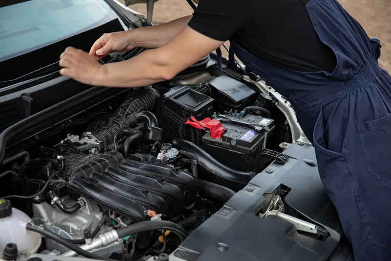 A mechanic in navy overalls leaning over an open bonnet, checking the engine bay and battery.
